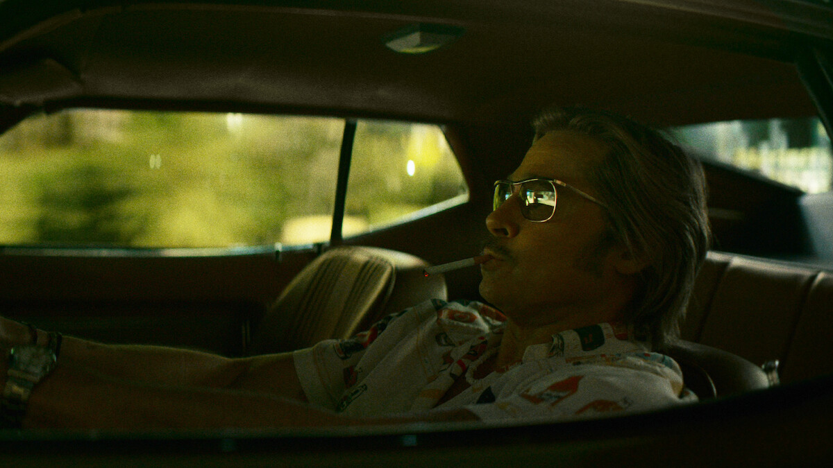 Brad Pitt como Cliff Booth, de óculos escuros, sentado em um carro.